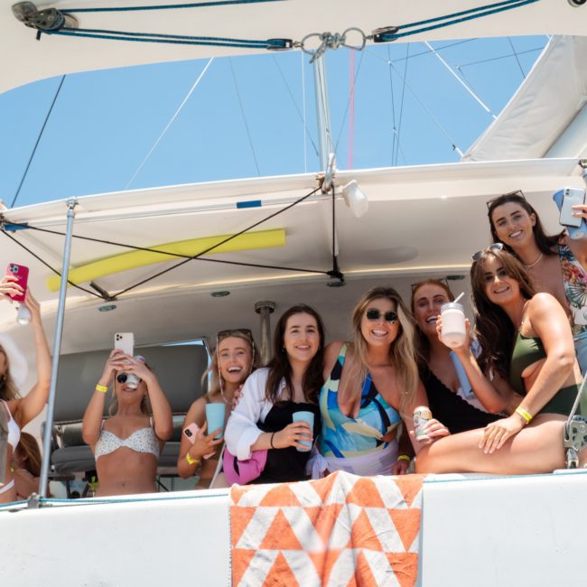 A group of eight people, mostly women, are on a boat. Some are holding drinks, and a few are using their phones. They are smiling and enjoying sunny weather on a private yacht charter in Sydney Harbour.