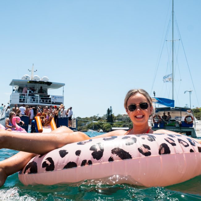 A person wearing sunglasses, a wristband, and a swimsuit floats on a leopard-print inflatable ring in the water, with boats and people in the background. Perfect for capturing the vibe of a Sydney boat party hire or indulging in luxury yacht hire Sydney for an unforgettable experience.