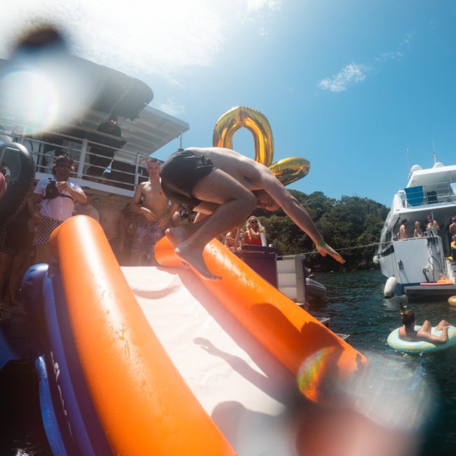 A person slides down an orange and white inflatable slide into the water, surrounded by other people and boats. Balloons and swimmers are visible in the background, creating a lively atmosphere perfect for a Catamaran party in Sydney.