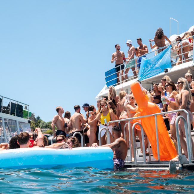 A lively gathering unfolds on a boat and swim platform, with some in swimsuits enjoying an inflatable pool. An orange inflatable figure adds to the fun. Ideal for a catamaran party Sydney or corporate boat events Sydney, this scene epitomizes joy on the water.