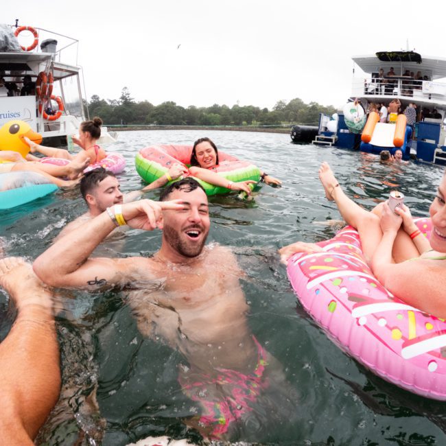 People floating on inflatables in the water next to a boat, enjoying themselves with one person making a peace sign. Other boats and people are visible in the background, making it an ideal setting for private yacht charters on Sydney Harbour.