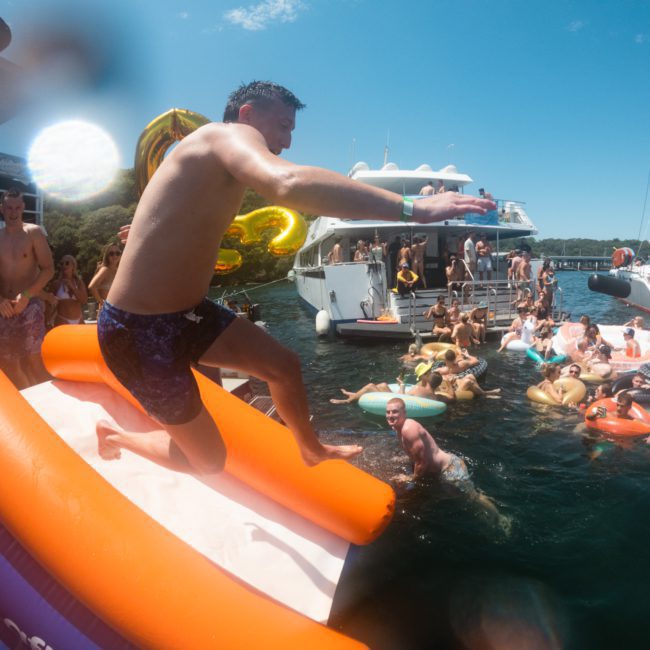 A man is mid-jump from an inflatable slide into a body of water surrounded by boats and people swimming and floating on inflatables, enjoying a festive Sydney boat party hire.