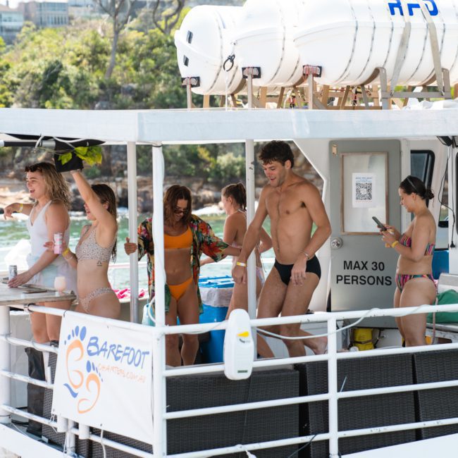 A group of people in swimwear are on a private yacht charter in Sydney Harbour, some dancing and some standing. Lifeboats are visible above them. The boat is by a wooded shoreline. A banner reads "BAREFOOT" with a footprint logo.