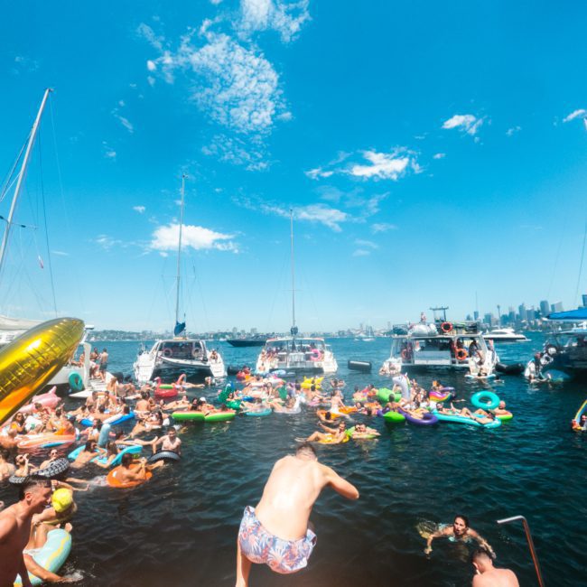 People enjoying a sunny day on a lake with inflatable floaties, yachts, and kayaks. A person is about to jump into the water from a private yacht charter in Sydney Harbour.