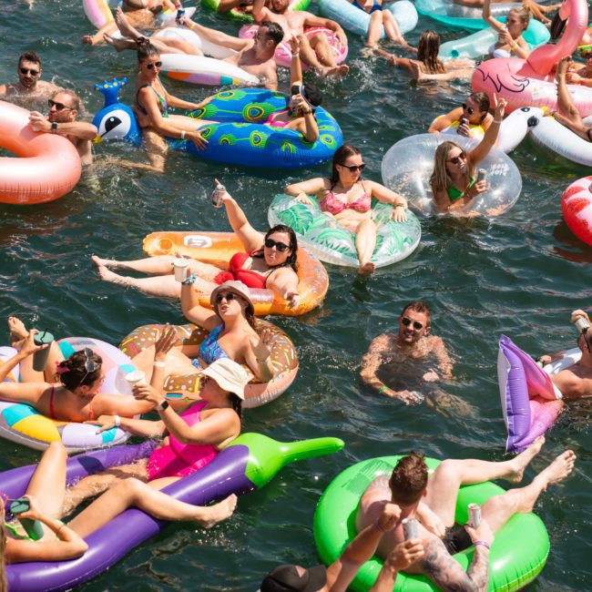A crowd of people enjoy a sunny day floating on colorful inflatable pool toys in a body of water, while a private yacht charter sails nearby.