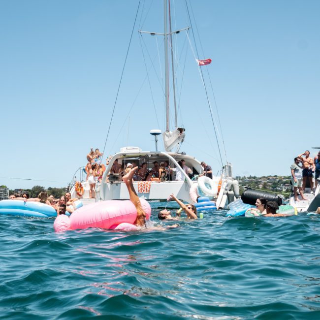 People enjoying a sunny day on the water, swimming and relaxing on inflatable floats near anchored boats during a luxury yacht hire Sydney event.