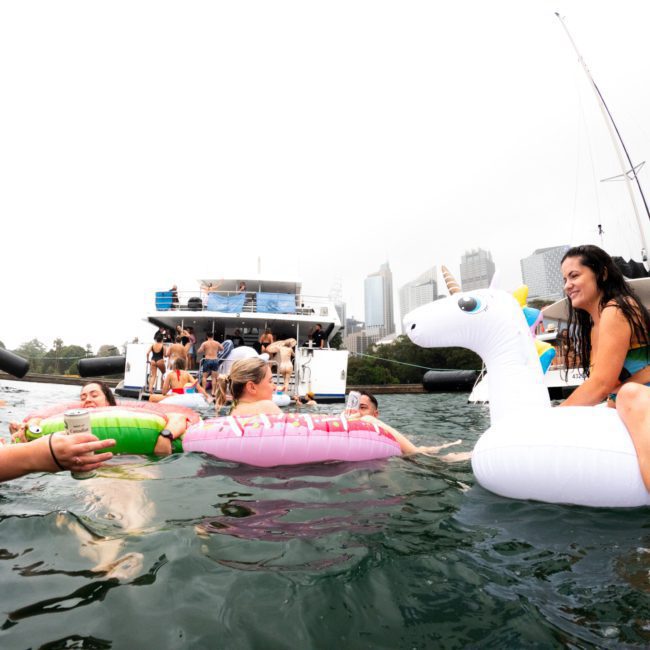 Group of people enjoying a catamaran party on inflatable floats in a body of water, with boats and city buildings in the background.