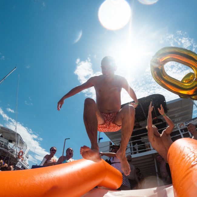 A person in shorts jumps from an inflatable structure under a sunny sky during a luxury yacht hire Sydney event, surrounded by people.