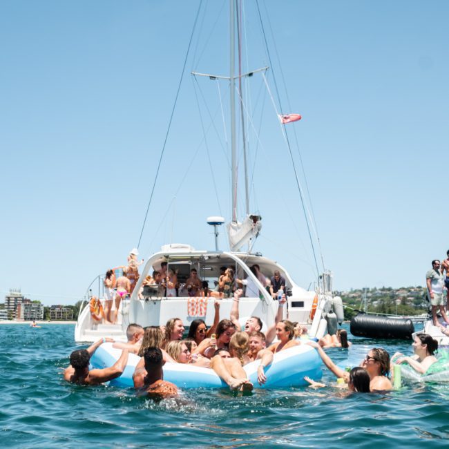 A large group of people are gathered on and around a luxury yacht, with some in the water using inflatables. The background shows a clear blue sky and buildings on the shoreline, exemplifying an exquisite private yacht charter in Sydney Harbour.