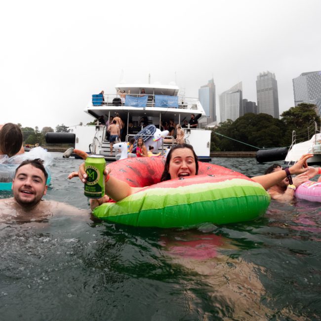 People are floating in the water with inflatable rings near boats. Two individuals in the foreground smile, one holding a can. City buildings are visible in the background, hinting at a lively catamaran party Sydney is famous for.
