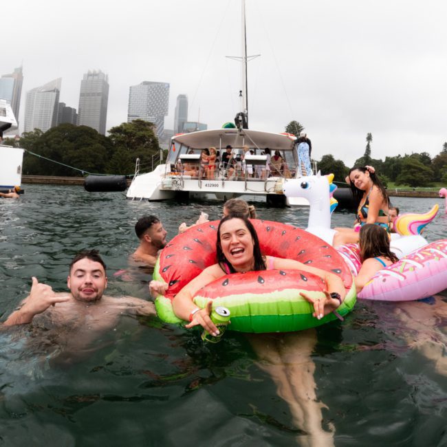 People swimming and floating on inflatable toys in a body of water, with boats and a city skyline in the background, all enjoying the perfect backdrop for a Corporate boat event Sydney.