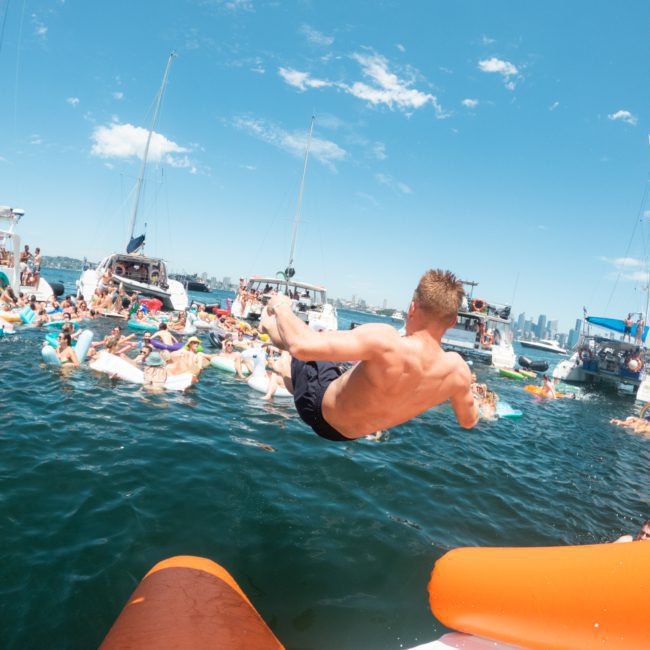 A shirtless man is captured mid-air as he jumps into a body of water surrounded by several boats and people on floaties, with a clear blue sky in the background. The vibrant scene evokes the vibe of luxury yacht hire in Sydney Harbour.