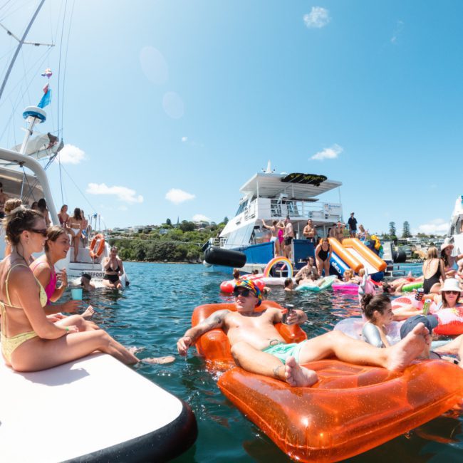 A group of people enjoy a sunny day on the water, relaxing on various inflatable floats and boats. Some individuals are sitting on the edge of a boat, while others float nearby. It's the perfect setting for a Sydney boat party hire experience.
