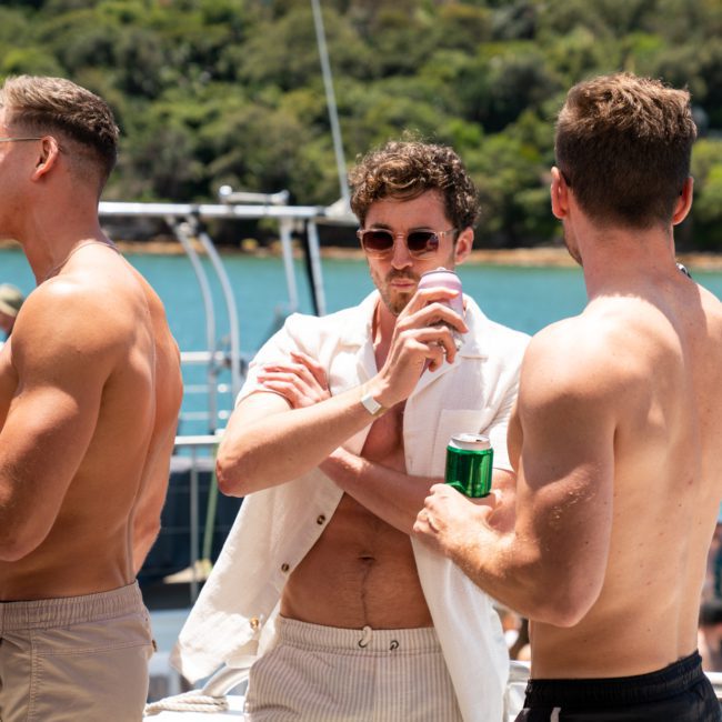 Three men in swimwear stand on a boat, each holding a drink, with lush greenery and water in the background. Perfect for your next DJ boat hire Sydney adventure.