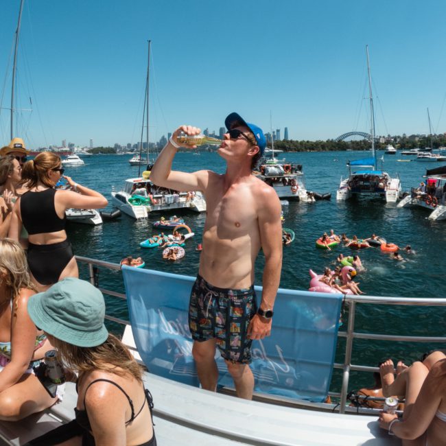 People on a boat enjoy a sunny day on the water. A man in a blue cap drinks from a bottle while others relax and socialize. In the background, several boats and people on inflatables can be seen, typical of a private yacht charter Sydney Harbour experience.