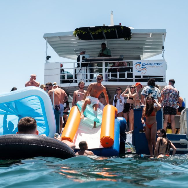 A group of people enjoying a sunny day on a catamaran with a slide and floating toys in the water. The boat's deck is filled with people socializing, making it perfect for corporate boat events Sydney.