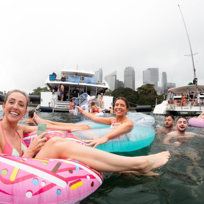 People in a city harbor on inflatable floats, holding drinks, with boats and skyscrapers in the background on a cloudy day. Consider Sydney boat party hire for your next gathering or corporate boat events Sydney to turn such scenes into unforgettable experiences.