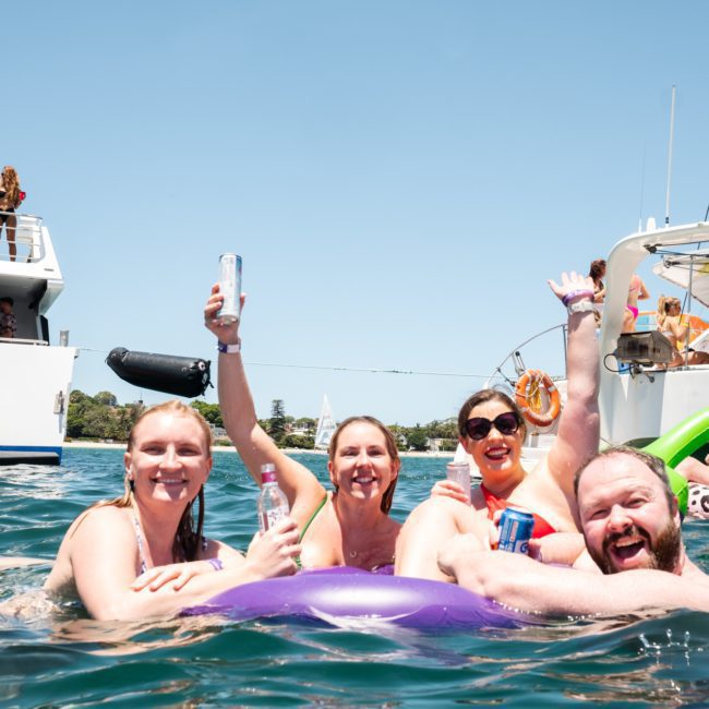 A group of four people enjoying themselves in the water near a boat, holding beverages and smiling. Other people are visible on private yacht charters in Sydney Harbour in the background.