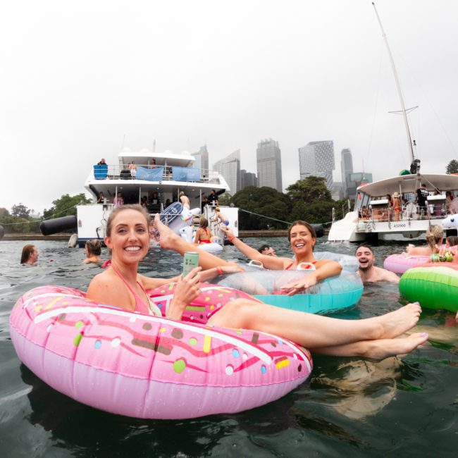 People on inflatable pool floats are enjoying a social gathering on the water near several boats with a city skyline in the background, featuring a remarkable DJ boat hire Sydney experience.