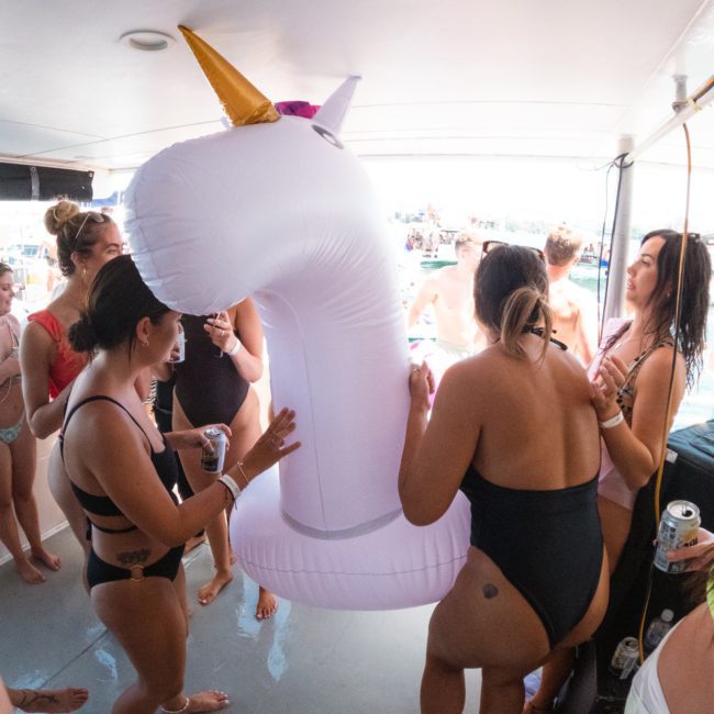 A group of people in swimwear gather on a private yacht charter in Sydney Harbour, interacting with a large inflatable unicorn.