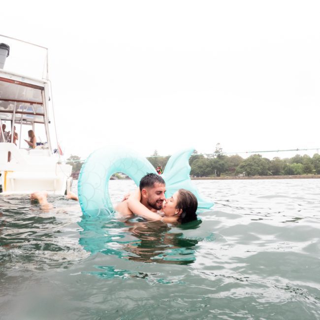 A couple embraces on a blue mermaid tail float in the water beside a boat, with trees in the background, reveling in their private yacht charter Sydney Harbour.
