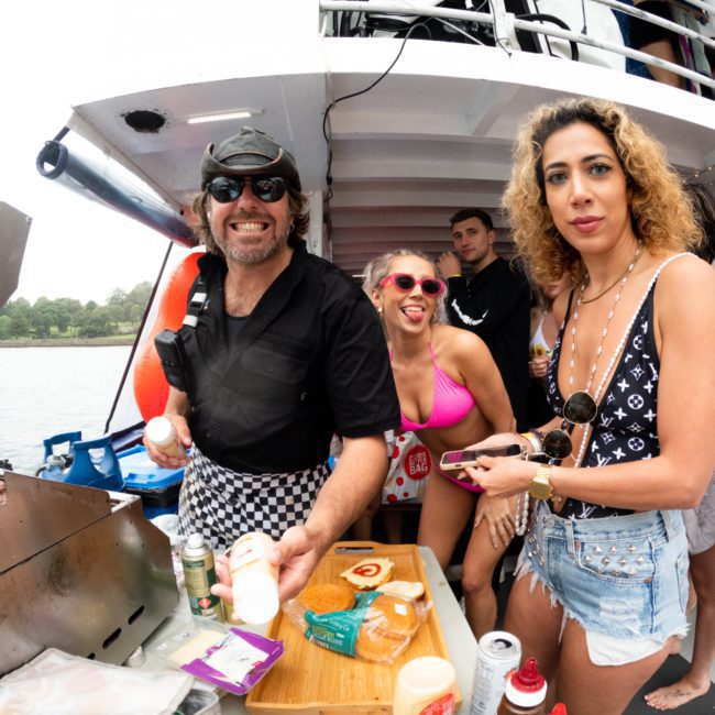 A group of people on a boat are gathered around a grill during a luxury yacht hire in Sydney. Food items are laid out on a wooden tray, and several of them are smiling and looking at the camera.