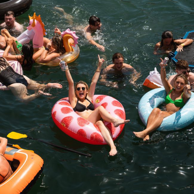 A group of people is enjoying a sunny day floating in a body of water, using various inflatables including a strawberry donut, unicorn, and circular floaties. Some are holding drinks and smiling, reminiscent of the fun you can have with a DJ boat hire Sydney.