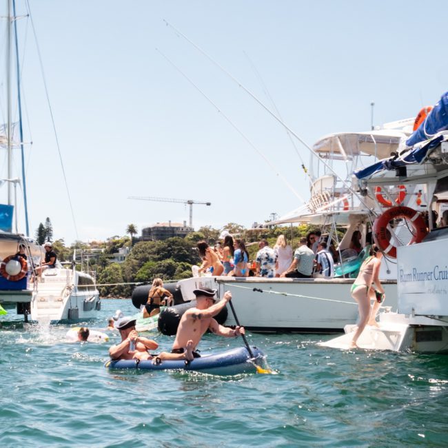 People enjoying water activities near boats anchored close to shore on a sunny day. Some are relaxing on inflatables while others prepare to dive or climb back onto the boats, perfect for a luxury yacht hire in Sydney.