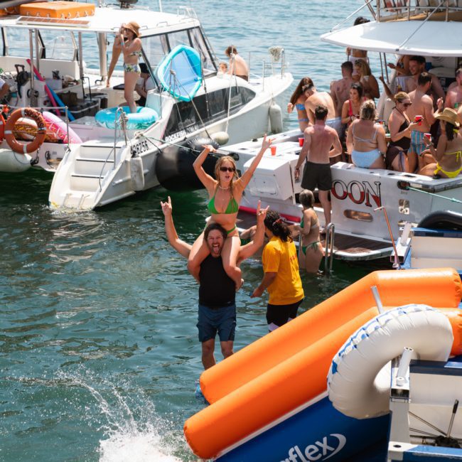 A group of people are enjoying a Sydney boat party hire on a sunny day. One person is being carried on another's shoulders while others socialize in and around the water. An inflatable slide is visible in the foreground, adding to the fun of the private yacht charter on Sydney Harbour.