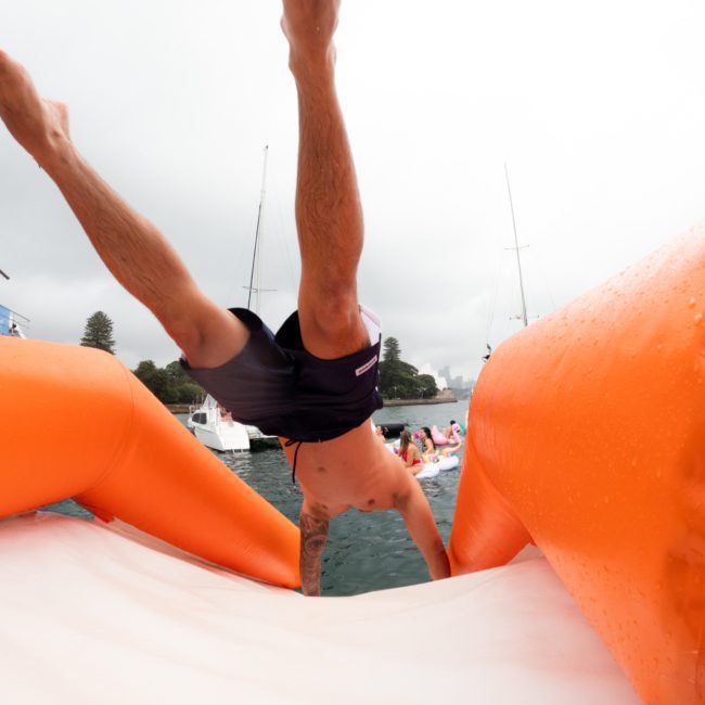 A person is seen mid-air after jumping from an orange inflatable structure into the water, with boats and other people visible in the background, enjoying a DJ boat hire Sydney.