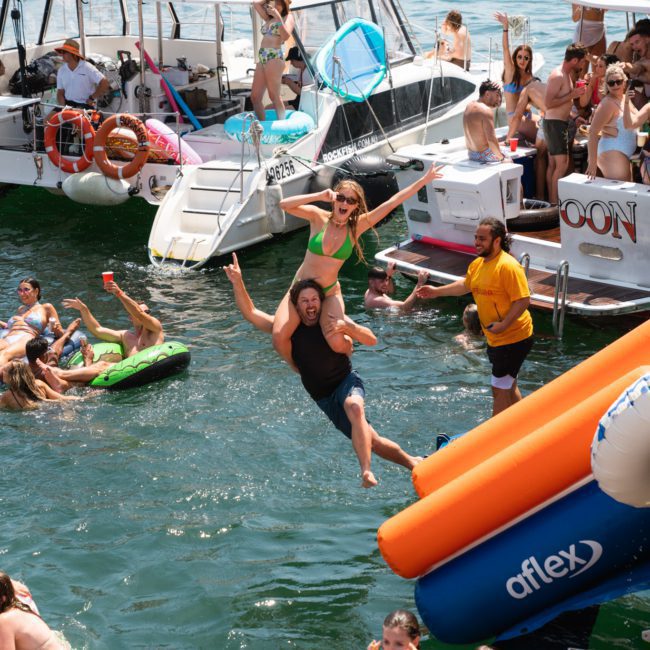 A lively scene with people enjoying a Sydney boat party hire in and around the water. A man is jumping into the water with a woman on his shoulders, while others socialize or swim nearby on a catamaran party in Sydney.