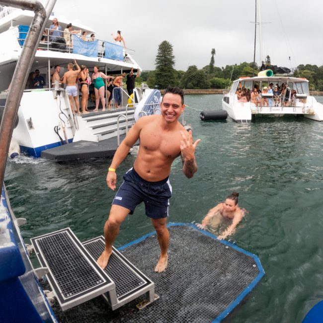A shirtless man poses while making a hand gesture on a luxury yacht hire Sydney. People are on the boat and in the water nearby, with another boat in the background. Trees and buildings are visible in the distance.