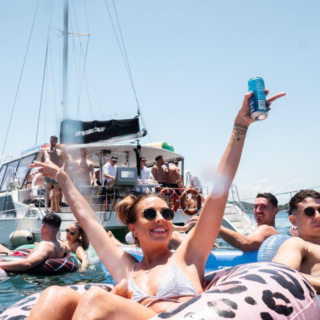 People enjoying a sunny day on a boat and in the water, with one person in the foreground holding a can and smiling. Various inflatable floats are visible around them, showcasing the fun of a Sydney boat party hire.