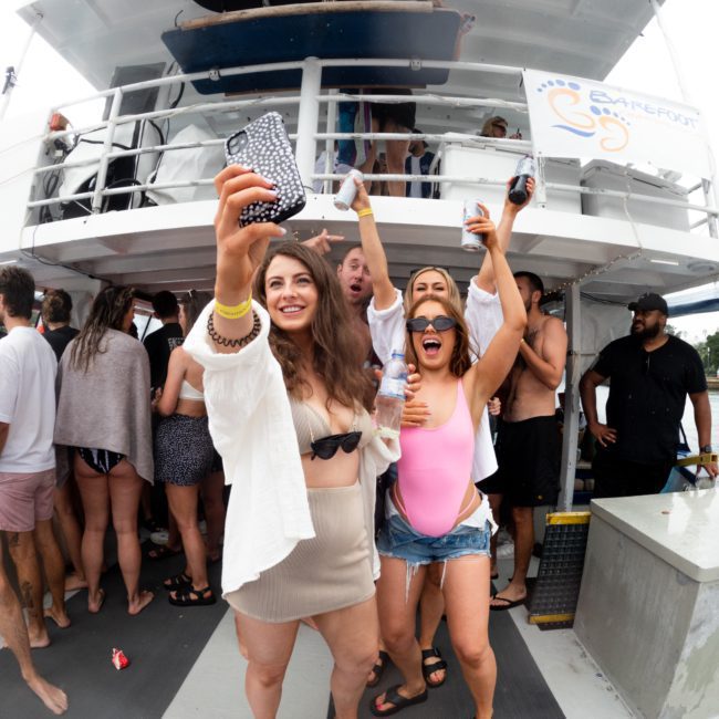 A group of people on a boat, with three women in the foreground smiling and posing for a selfie. One woman is holding up a phone, while others are socializing in the background at a lively Sydney boat party hire.