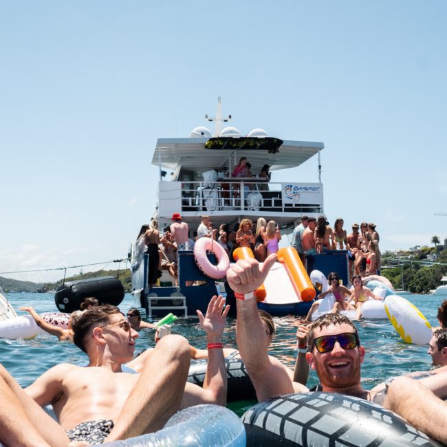 A group of people are floating on inflatable tubes in the water near a boat with more people on board, enjoying a private yacht charter in Sydney Harbour under a clear blue sky.