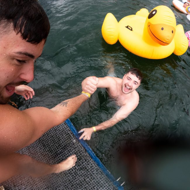 A man in the water gets help climbing onto a dock, surrounded by inflatable floaties, including a large yellow duck, during a private yacht charter on Sydney Harbour.