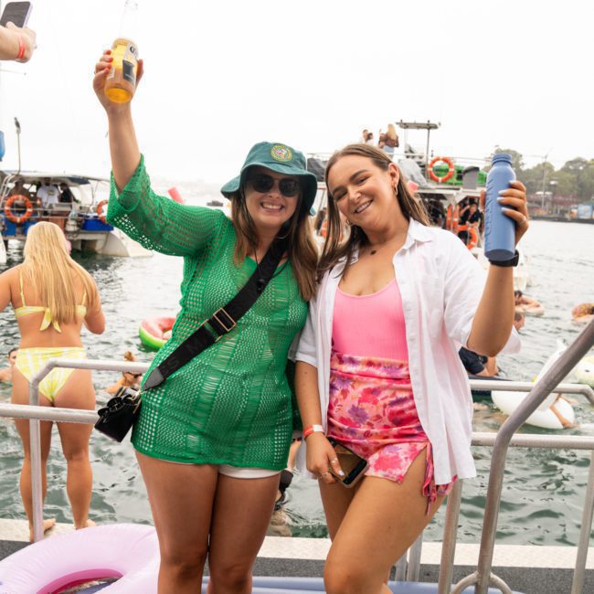 Two women are smiling and holding drinks on a dock by the water. One is wearing a green outfit with a hat, and the other is in a white shirt and pink shorts. People and boats, including a catamaran party Sydney-style, are visible in the background.