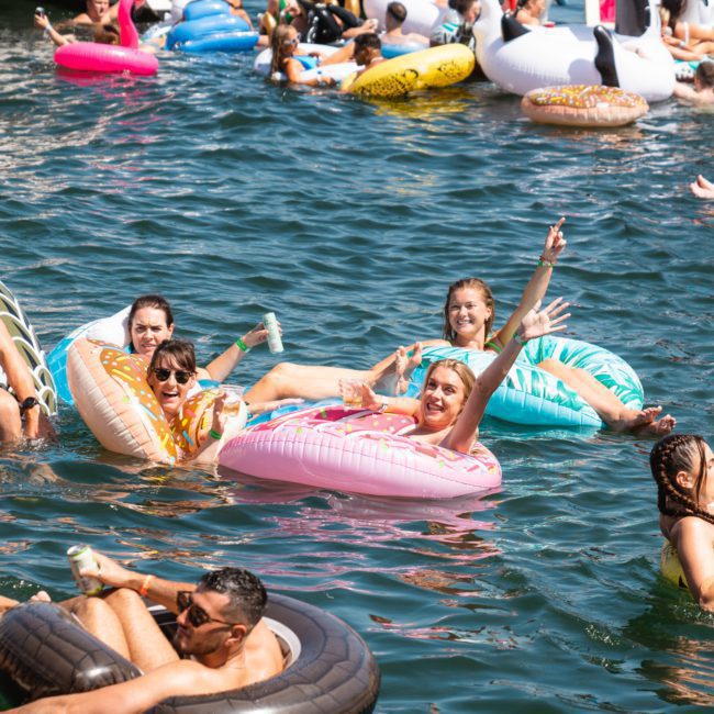A group of people enjoy a sunny day floating on inflatable pool toys in a body of water, holding drinks and socializing during a lively Sydney boat party hire.