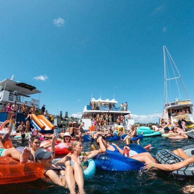 A large group of people enjoying a sunny day in the water, floating on various inflatables near boats anchored close together, reveling in a vibrant Sydney boat party hire.