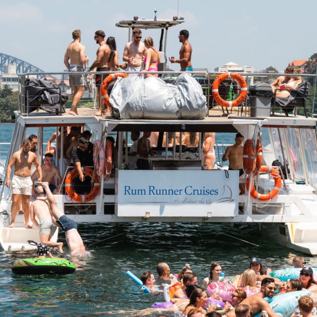 A group of people are enjoying a sunny day on a boat and in the water, labeled "Rum Runner Cruises," with a cityscape and bridge in the background. Some are on inflatables, and others are on the boat's deck. It's an ideal scene for luxury yacht hire Sydney enthusiasts.