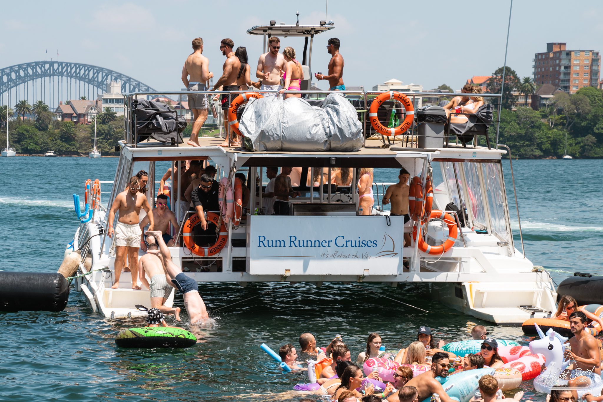 A group of people enjoying a corporate boat event with inflatables in the water on a sunny day. The boat has a "Rum Runner Cruises" sign. Sydney Harbour Bridge is visible in the background.