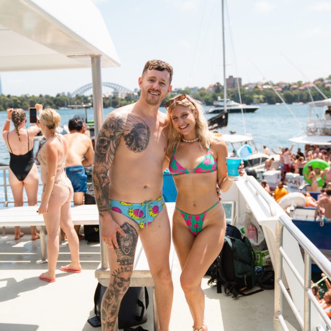A man and a woman in swimsuits stand on a boat deck with other people, enjoying the scenic cityscape in the background, perfect for corporate boat events Sydney.