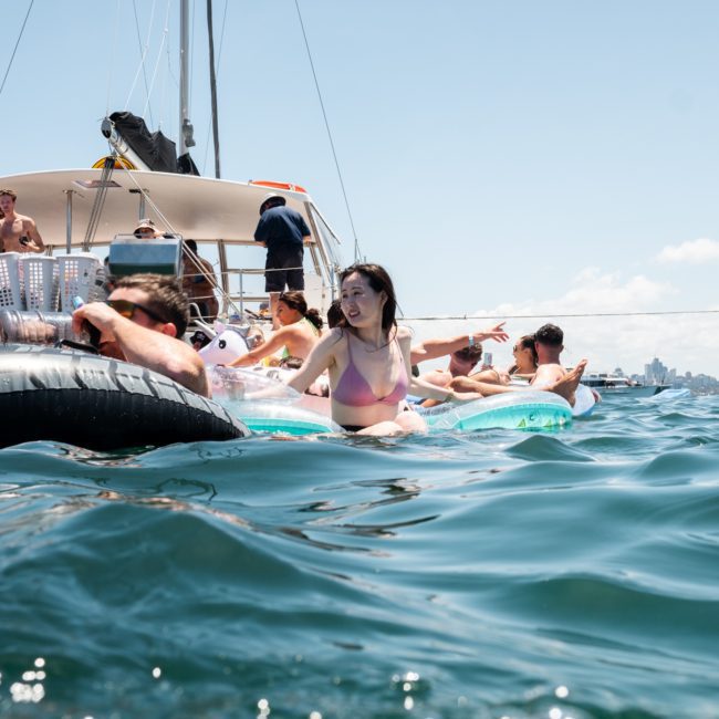 People are floating on inflatable tubes in the water near a catamaran party in Sydney on a sunny day. Some are sitting on the boat while others are enjoying the water.