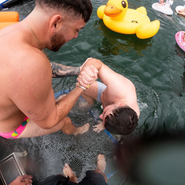 A man helps another man out of the water onto a platform. Several people on inflatable floats are visible in the background, enjoying the day near a private yacht charter on Sydney Harbour.