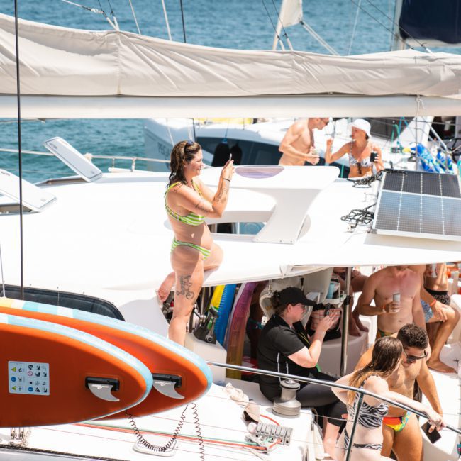 Many people are gathered on a yacht in swimwear, enjoying the sun. A woman on the upper deck is capturing memories with her phone. An orange paddleboard is mounted on the side of this luxury yacht hire Sydney, making it a perfect scene for any catamaran party Sydney.