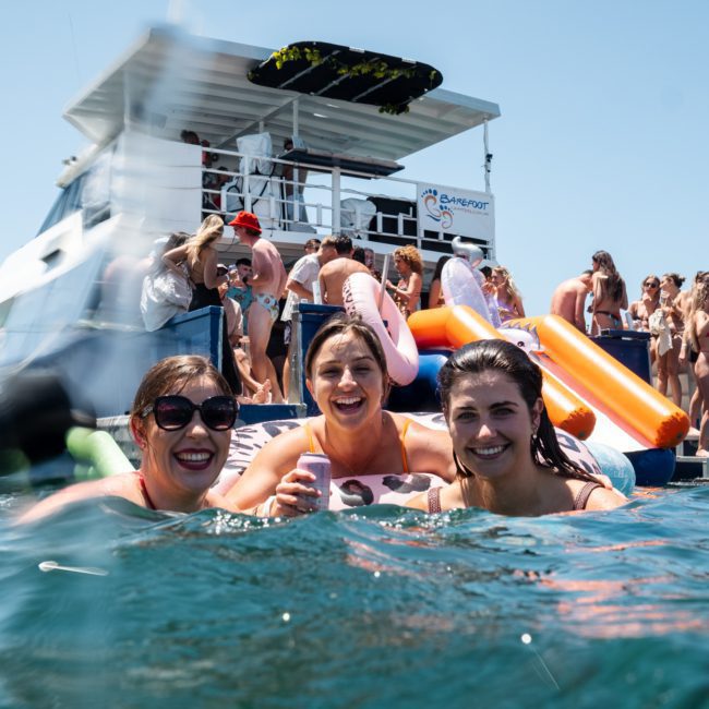 Three people smiling while swimming in the water, with a group of people on a boat in the background. The scene is set on a sunny day, perfect for a private yacht charter Sydney Harbour.