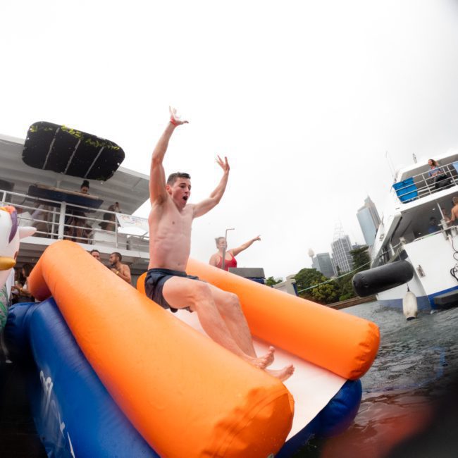 A person slides down an inflatable slide into the water from a luxurious yacht, surrounded by other participants and boats. It's the perfect scene for a Sydney boat party hire on Sydney Harbour.
