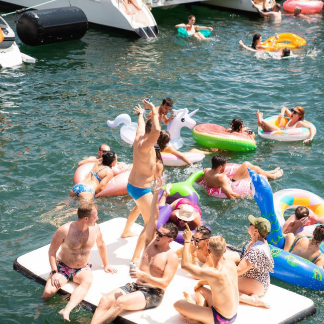 A group of people in swimsuits enjoy floating on inflatable devices and platforms in a body of water, with boats in the background. Some individuals are socializing while others relax on the inflatables, reminiscent of a lively catamaran party Sydney style or private yacht charter Sydney Harbour event.
