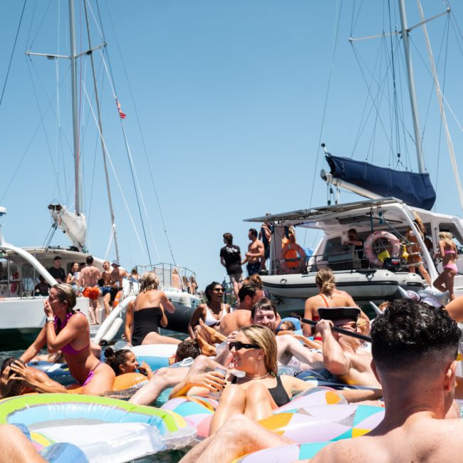 A group of people on inflatable floats enjoy a sunny day in the ocean near several sailboats, setting the perfect scene for a catamaran party in Sydney.