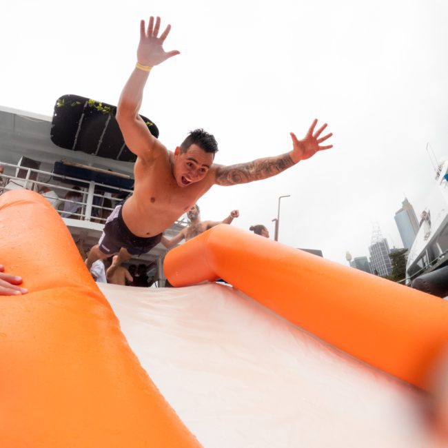 A man is joyfully sliding down an orange inflatable slide on a boat, with arms raised and an excited expression, under a cloudy sky. Other people and boats are visible in the background, enjoying what seems to be a lively Sydney boat party hire.